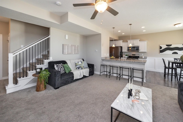 living area with a ceiling fan, stairway, dark colored carpet, a decorative wall, and recessed lighting