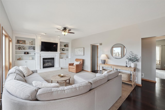 living area featuring built in features, a glass covered fireplace, wood finished floors, baseboards, and ceiling fan