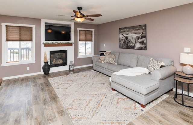 living room featuring a ceiling fan, a large fireplace, baseboards, and wood finished floors