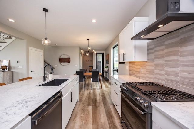 kitchen featuring stainless steel appliances, a sink, white cabinets, wall chimney exhaust hood, and decorative light fixtures