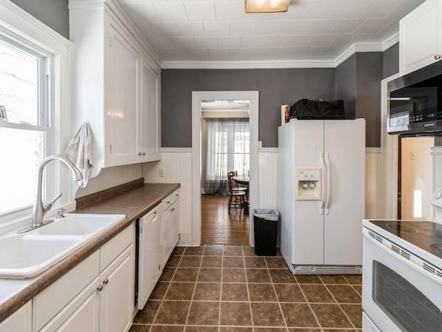 kitchen with white appliances, a wainscoted wall, crown molding, white cabinetry, and a sink