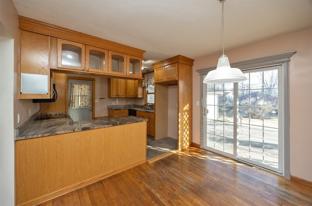 kitchen with a peninsula, wood finished floors, brown cabinets, dark countertops, and glass insert cabinets