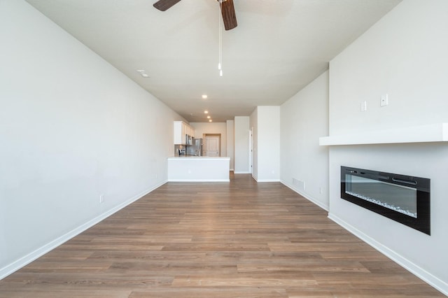 unfurnished living room featuring ceiling fan, wood finished floors, visible vents, baseboards, and a glass covered fireplace