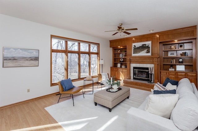 living area with light wood finished floors, a tiled fireplace, baseboards, built in shelves, and a ceiling fan