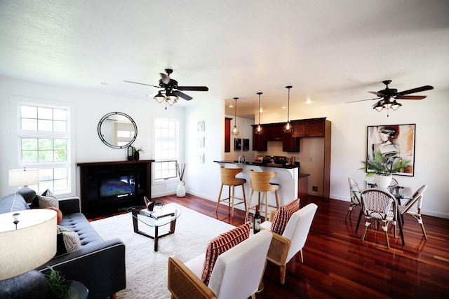 living area with a ceiling fan, dark wood-type flooring, a wealth of natural light, and a glass covered fireplace