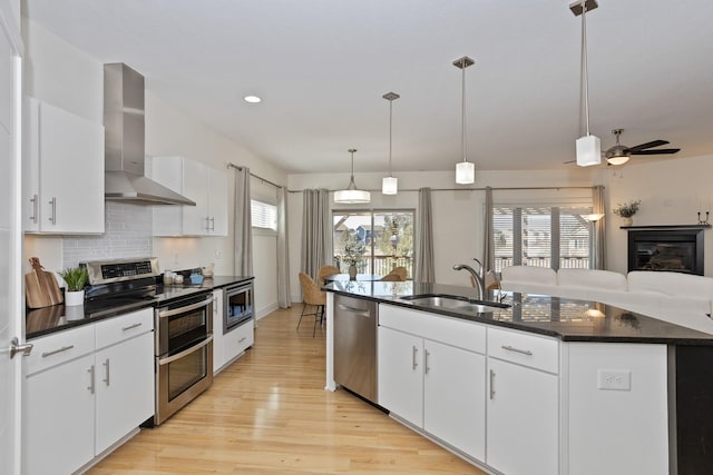 kitchen featuring stainless steel appliances, a sink, wall chimney range hood, light wood-type flooring, and tasteful backsplash