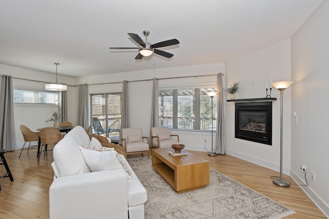 living area with light wood finished floors, a glass covered fireplace, a ceiling fan, and baseboards