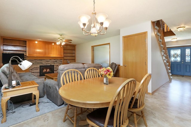 dining room featuring concrete flooring, stairway, a stone fireplace, and ceiling fan with notable chandelier
