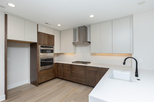 kitchen with wall chimney exhaust hood, appliances with stainless steel finishes, light countertops, light wood-type flooring, and a sink