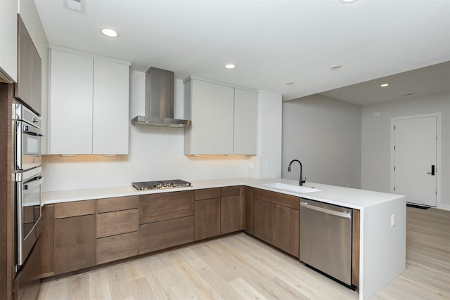 kitchen featuring stainless steel appliances, a sink, wall chimney range hood, modern cabinets, and a peninsula