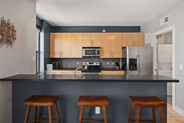 kitchen with appliances with stainless steel finishes, stacked washer / dryer, backsplash, and a kitchen breakfast bar