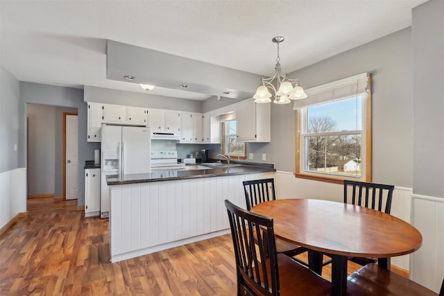 kitchen featuring under cabinet range hood, a peninsula, white appliances, light wood finished floors, and dark countertops