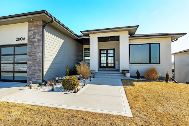 view of exterior entry featuring a garage, a lawn, stone siding, french doors, and stucco siding