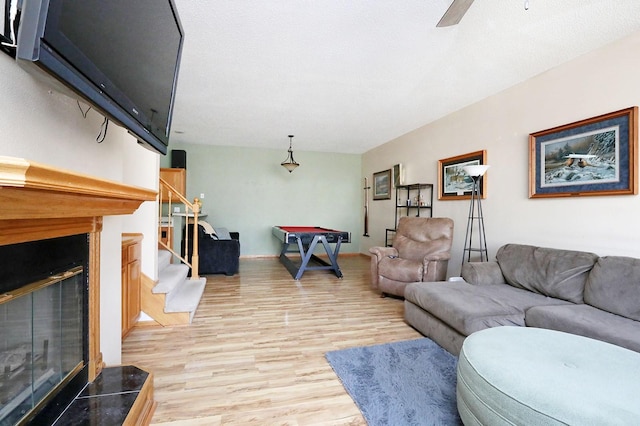 living area featuring stairway, a glass covered fireplace, light wood-style flooring, and a ceiling fan