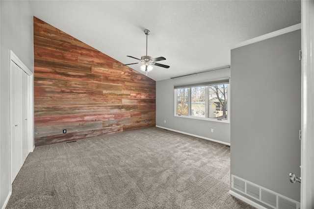 carpeted empty room with a textured ceiling, wooden walls, a ceiling fan, visible vents, and vaulted ceiling