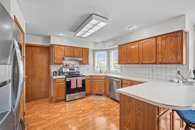 kitchen featuring a peninsula, under cabinet range hood, brown cabinets, stainless steel appliances, and a sink