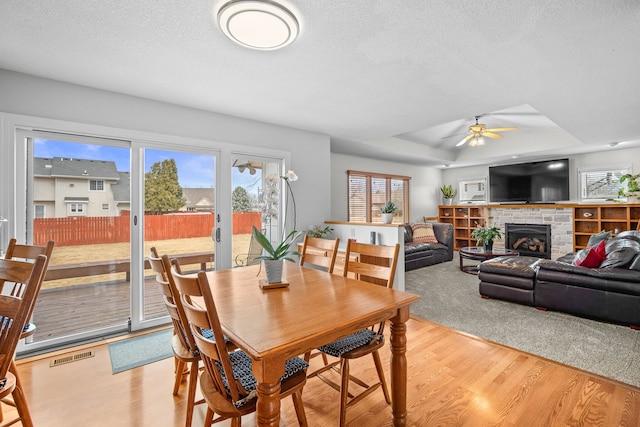 dining room featuring a fireplace, a tray ceiling, light wood-type flooring, a ceiling fan, and visible vents