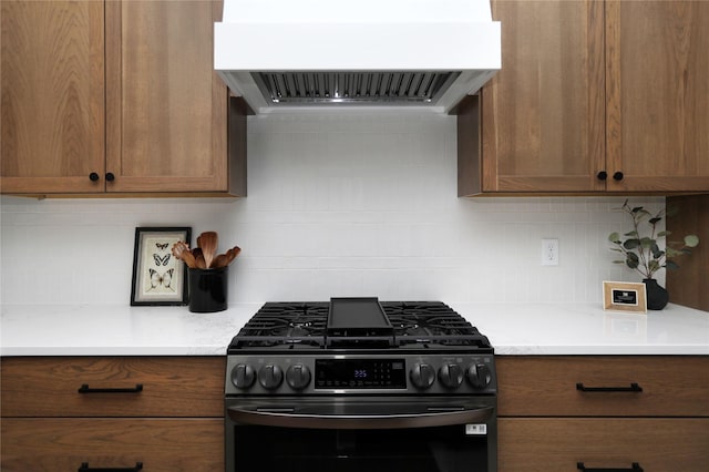 kitchen with brown cabinets, light countertops, backsplash, black gas range, and exhaust hood