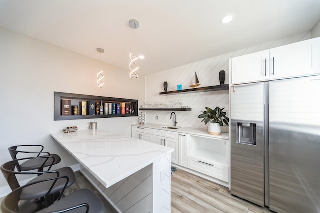 kitchen with a sink, white cabinetry, stainless steel fridge with ice dispenser, open shelves, and a kitchen bar