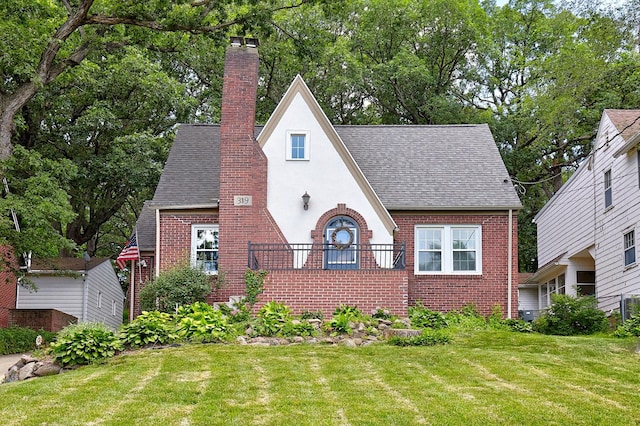 tudor house featuring stucco siding, a front yard, a shingled roof, brick siding, and a chimney