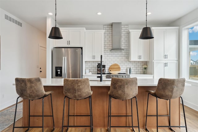 kitchen with light wood finished floors, a center island with sink, visible vents, stainless steel fridge with ice dispenser, and wall chimney range hood