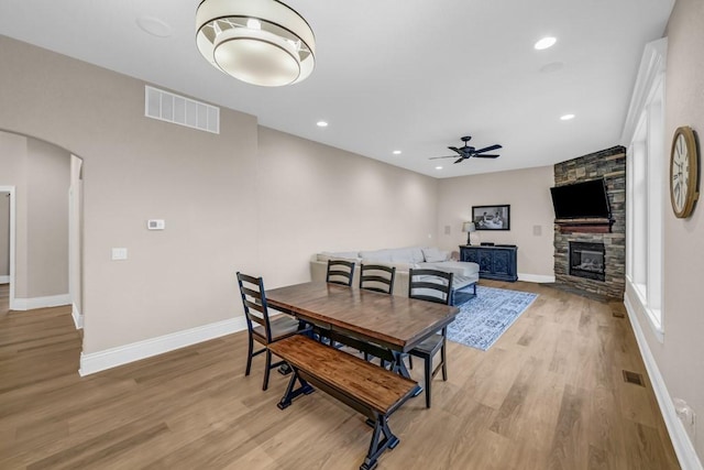 dining area with arched walkways, light wood finished floors, visible vents, ceiling fan, and a stone fireplace