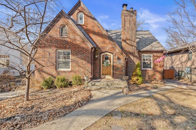 english style home featuring brick siding, a shingled roof, a chimney, and fence