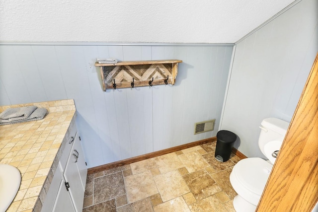 half bath featuring a textured ceiling, toilet, stone tile floors, vanity, and visible vents