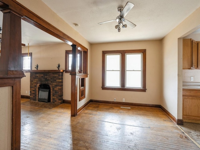 unfurnished living room featuring a ceiling fan, a textured ceiling, light wood-style floors, baseboards, and a brick fireplace