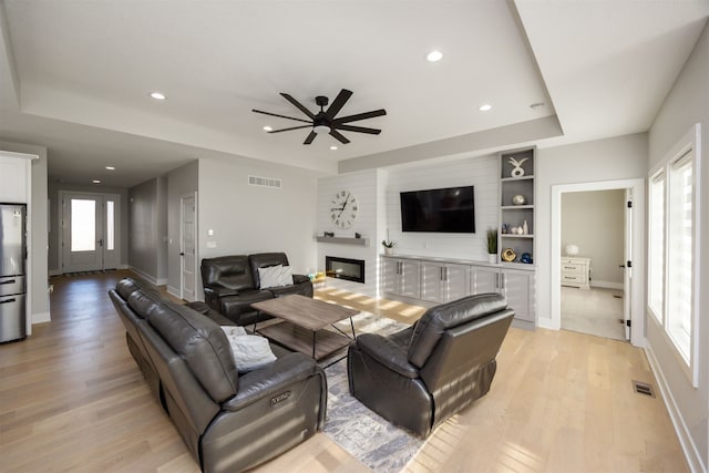 living room featuring visible vents, a raised ceiling, light wood-style floors, and a glass covered fireplace