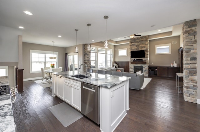 kitchen with an island with sink, light stone counters, a stone fireplace, stainless steel dishwasher, and a sink