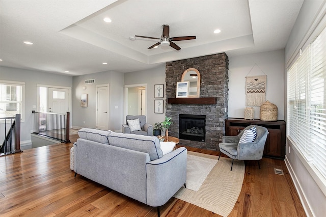 living room with a stone fireplace, a healthy amount of sunlight, a raised ceiling, and wood finished floors