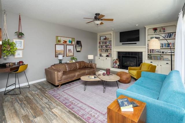 living room featuring built in shelves, wood finished floors, baseboards, ceiling fan, and a tiled fireplace