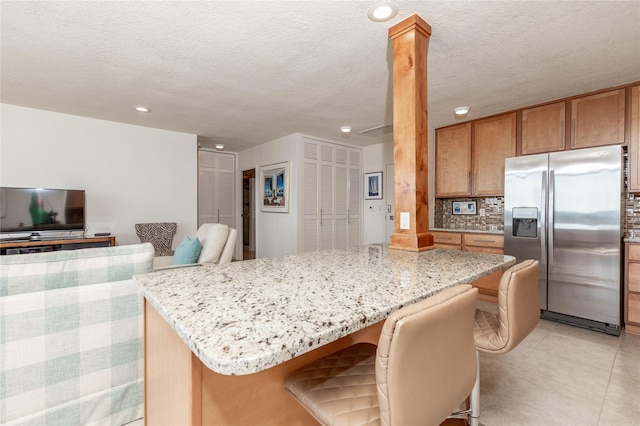 kitchen featuring light stone counters, brown cabinetry, stainless steel refrigerator with ice dispenser, a kitchen breakfast bar, and tasteful backsplash