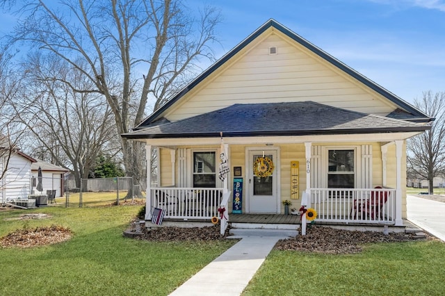 bungalow featuring a porch, fence, a front yard, a shingled roof, and central AC unit