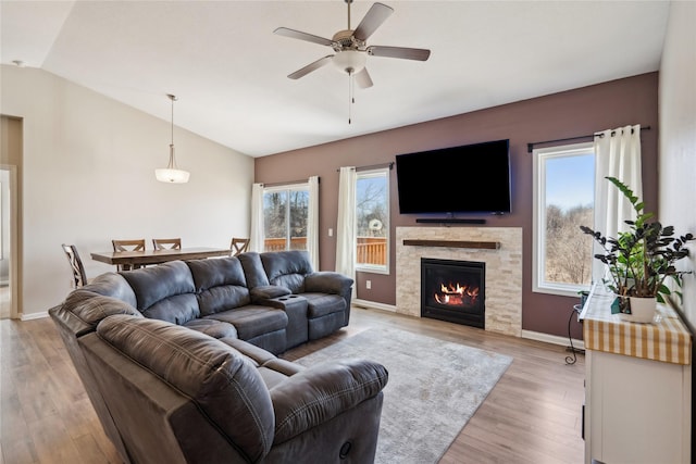 living area with a ceiling fan, baseboards, light wood-style flooring, a stone fireplace, and vaulted ceiling