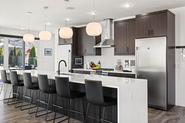 kitchen featuring a kitchen island with sink, stainless steel appliances, light countertops, dark brown cabinetry, and wall chimney exhaust hood