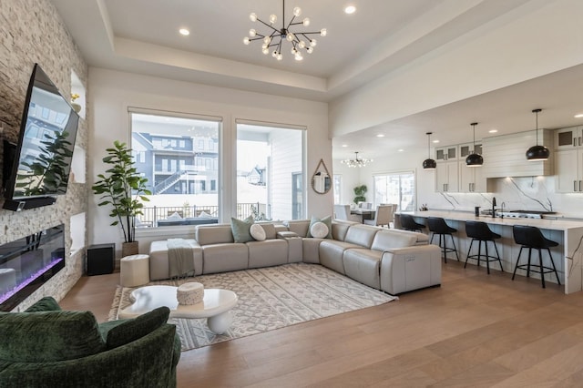 living room featuring an inviting chandelier, a tray ceiling, recessed lighting, a stone fireplace, and light wood-style floors