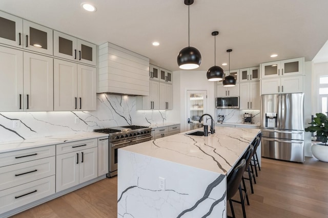 kitchen featuring light wood finished floors, a center island with sink, appliances with stainless steel finishes, custom exhaust hood, and a sink