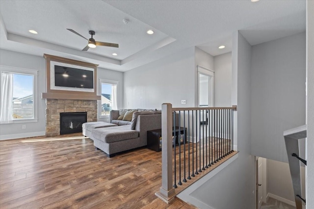 living area with a raised ceiling, wood finished floors, recessed lighting, a stone fireplace, and baseboards