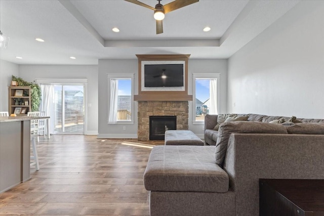 living room featuring a tray ceiling, a stone fireplace, wood finished floors, and a healthy amount of sunlight