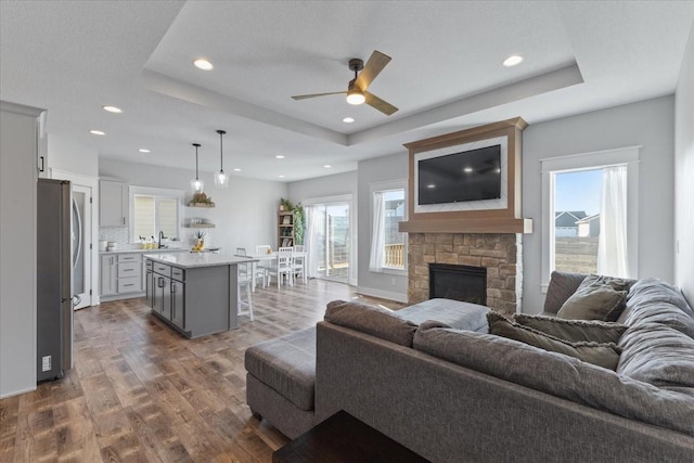 living room with dark wood-style floors, a stone fireplace, recessed lighting, and a tray ceiling