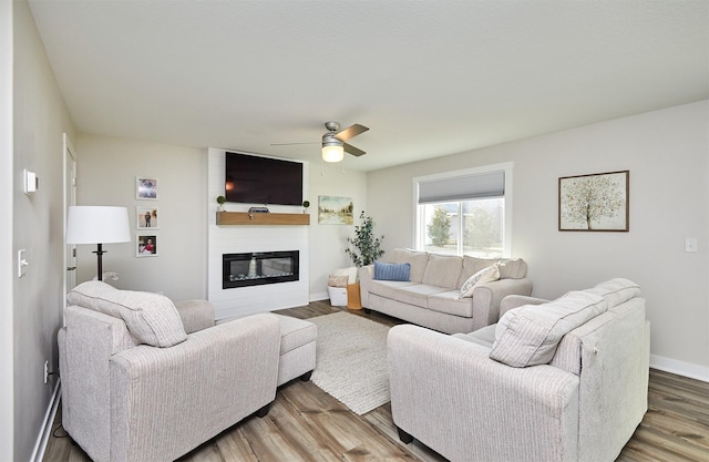 living area with a glass covered fireplace, baseboards, wood finished floors, and ceiling fan