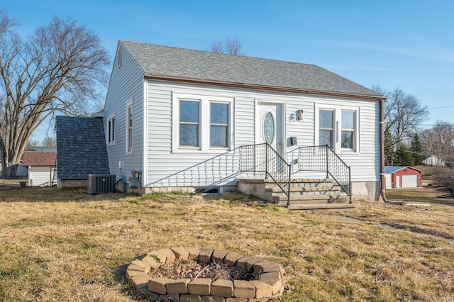 bungalow featuring a front lawn, central AC, a fire pit, roof with shingles, and a garage