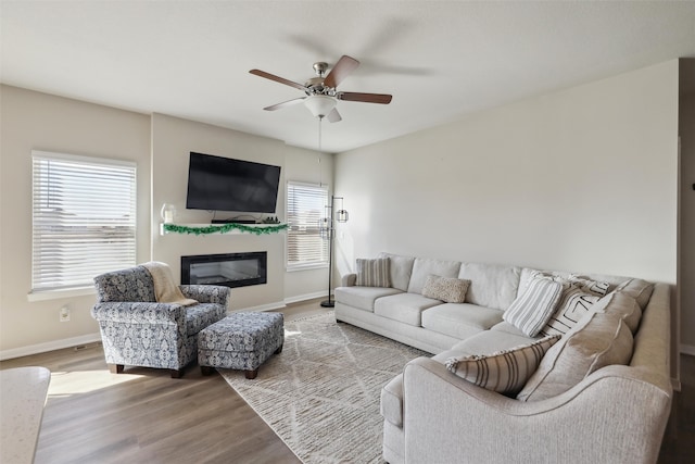 living area featuring a glass covered fireplace, baseboards, a ceiling fan, and wood finished floors