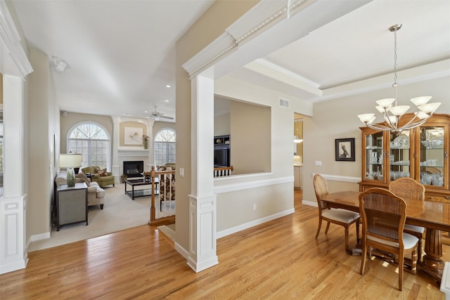 dining room with a tray ceiling, light wood-style flooring, decorative columns, a fireplace, and ceiling fan with notable chandelier