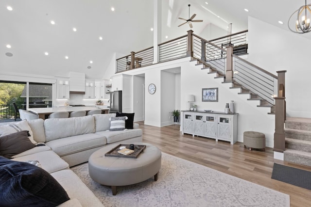 living room with baseboards, stairway, light wood-style flooring, ceiling fan with notable chandelier, and high vaulted ceiling