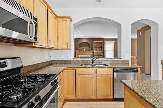 kitchen featuring light wood finished floors, light brown cabinetry, dark stone countertops, stainless steel appliances, and a sink