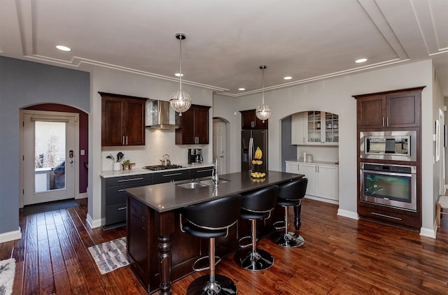 kitchen featuring a sink, dark wood finished floors, stainless steel appliances, arched walkways, and wall chimney range hood