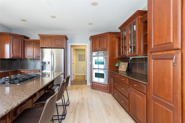 kitchen with dark stone countertops, a kitchen breakfast bar, stainless steel appliances, light wood finished floors, and glass insert cabinets